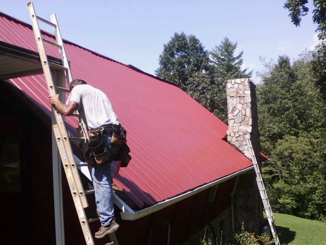 Roofer on a ladder beside a red metal roof and stone chimney — Soffit & Fascia Repair by Roofing Brothers in Molino